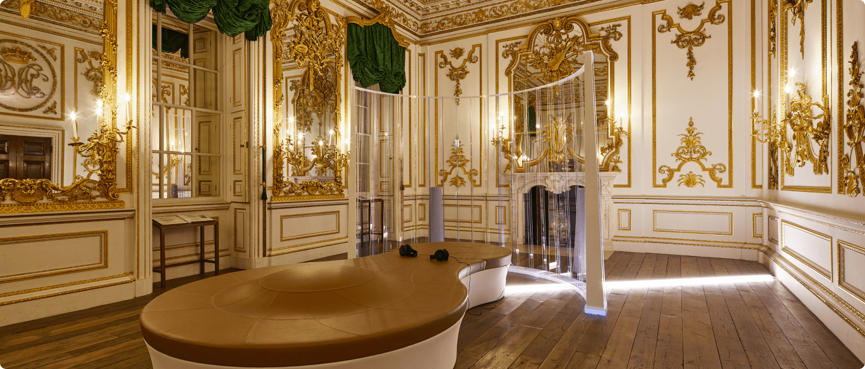 A gilded room at the V&A with white and gold panelling, a curved screen of vertical lit strands around a low leather bench with two pairs of headphones on it.