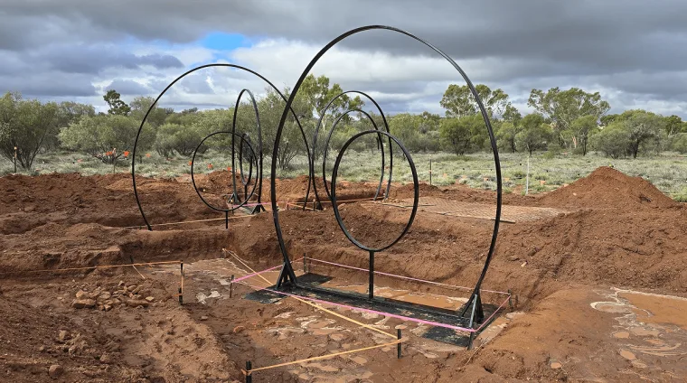Bare steel hoop frames standing in the red desert soil during the build, scrub and cloud behind them