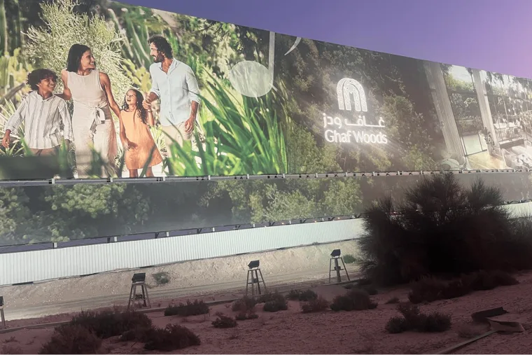 A large branded hoarding showing a family among greenery, floodlights standing on the bare ground in front of it