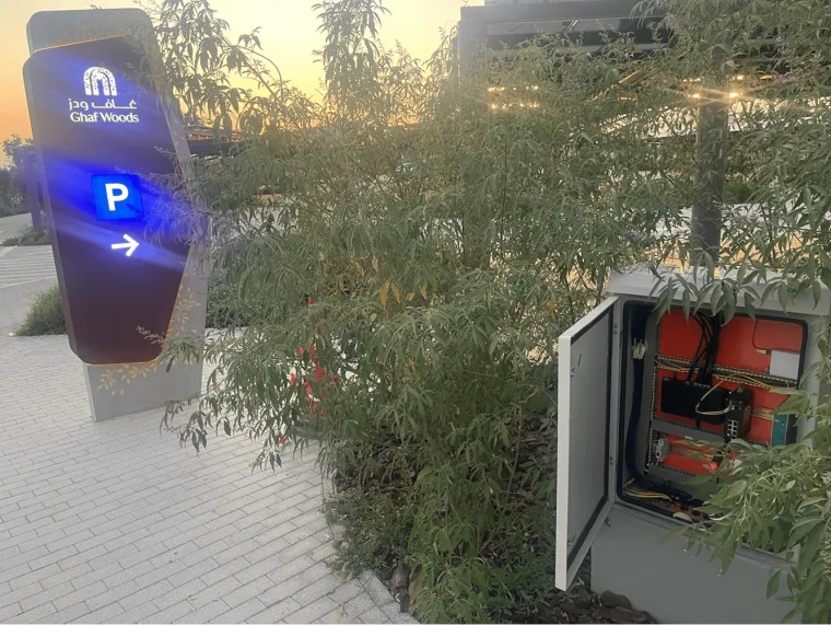 An illuminated wayfinding totem with a blue parking arrow at sunset, an open electrical enclosure among the shrubs beside it
