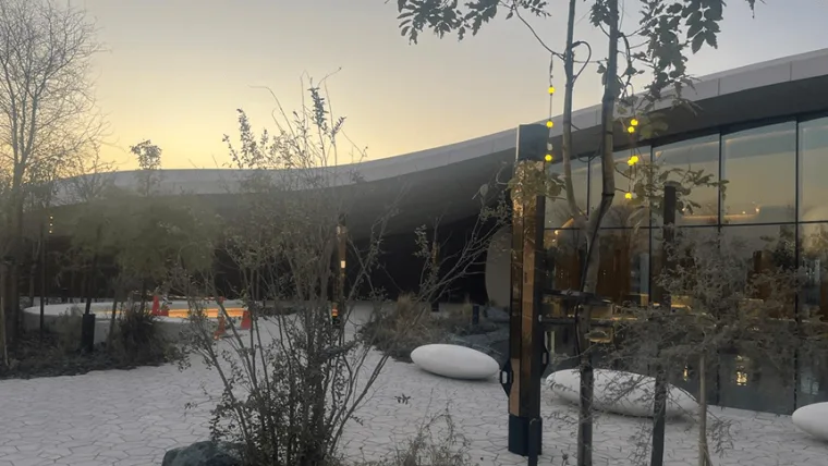 Dusk over the glass fronted pavilion, warm light poles standing among the planting and pale paving in front