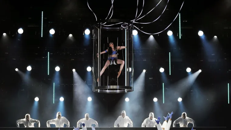 The tank flown above the stage with a dancer curled inside, a row of white-suited figures seated below in haze and spotlight beams