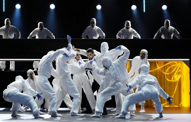 White-suited dancers piled into a scrum downstage, grey-suited figures watching from a level above, a yellow-lit panel to one side