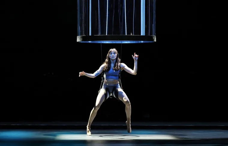 A solo dancer in blue body paint braced under a hanging ring trailing strands of blue light