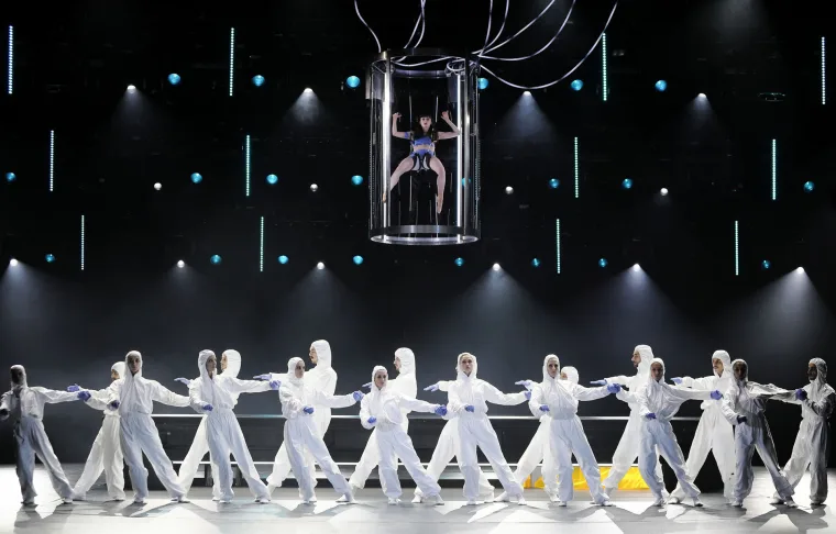 The full company in white suits linked hand to hand across the stage, the lit tank suspended above them