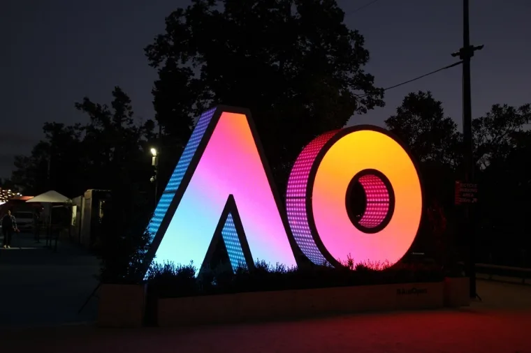 The sculpture seen from further down the path after dark, pink and orange faces glowing beside event marquees
