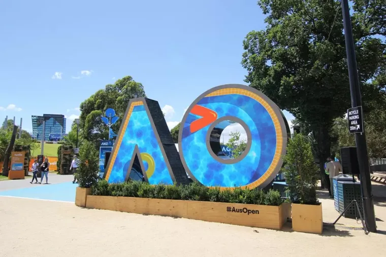 Blue mosaic and orange arrow artwork under a clear sky, visitors and event signage on the sand-coloured concourse