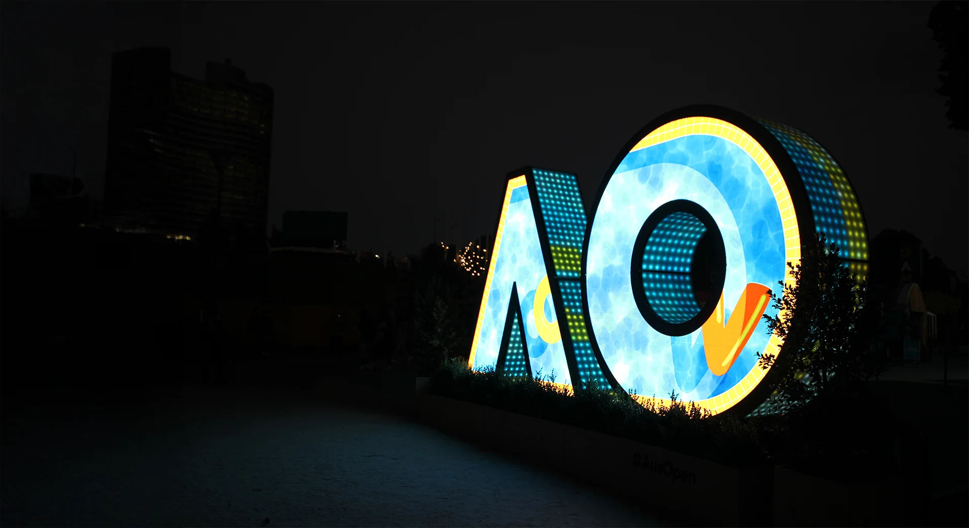 Wide night view of the Australian Open AO sculpture, blue water and yellow ring artwork across the letters and the darkened city skyline to the left