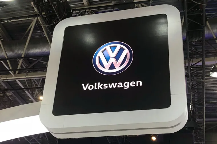 Illuminated Volkswagen logo sign on a white rounded frame, shot from below against the exhibition hall rigging