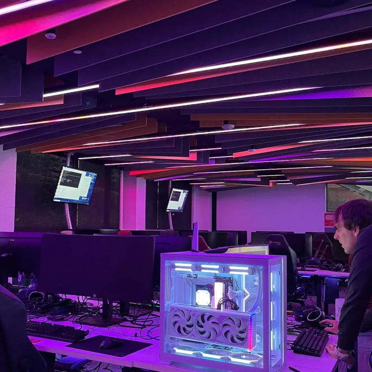A student at a keyboard on the right, a glass sided PC lit white in the foreground and magenta light across the ceiling