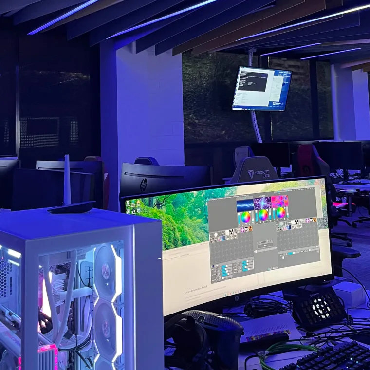 A gaming PC with a glowing white case beside an ultrawide monitor running ELM, colour wheels and effect thumbnails on screen, the darkened classroom behind