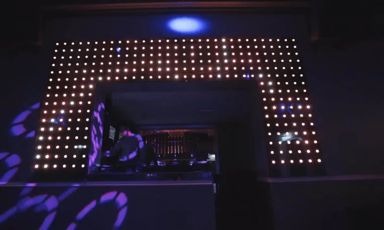 The DJ booth framed by a grid of warm white pixel dots, purple patterns thrown across the floor in front