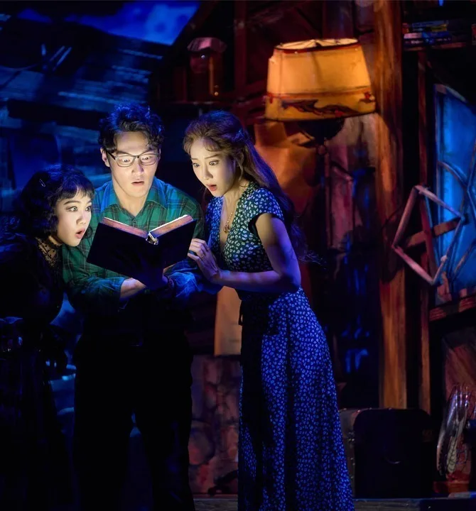 Three characters huddled over an open book in warm lamplight, a cluttered attic of beams and junk behind them