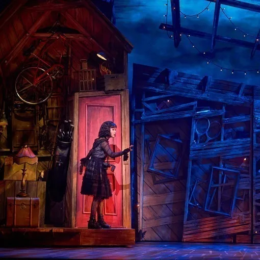A figure in black standing in a red-lit doorway, the attic around her washed cold blue