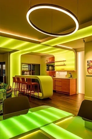 Kitchen and bar washed acid yellow and green, a ring pendant overhead and a lit dining table in the foreground