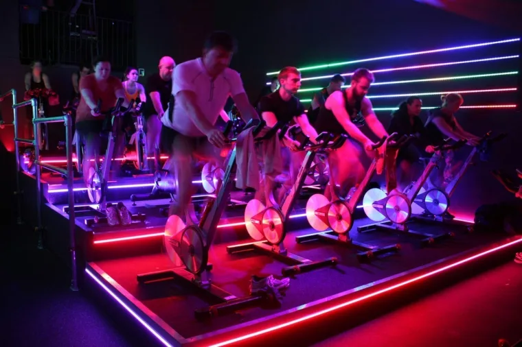 Rows of spin bikes with riders working, rainbow strips fanning across the back wall and a red line along the floor