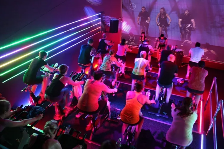 The studio from the back corner, a full class riding beneath the rainbow strips with a screen of cyclists on the far wall