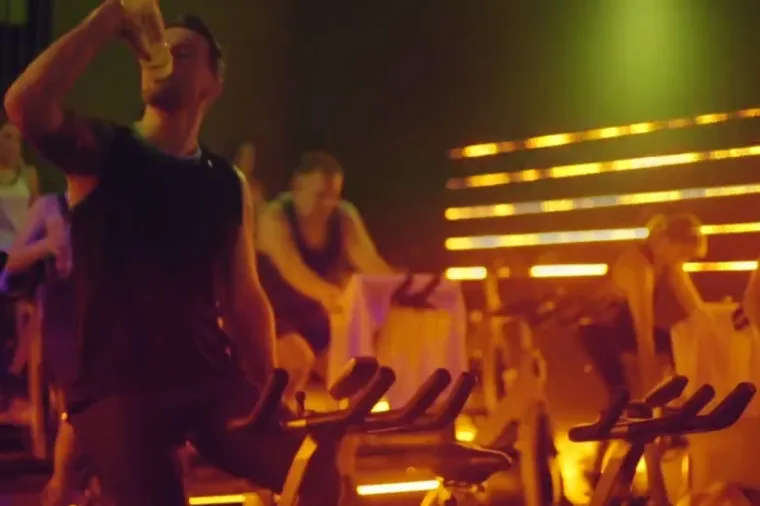 The class under amber and yellow strips, one man sitting up to drink from his bottle mid ride