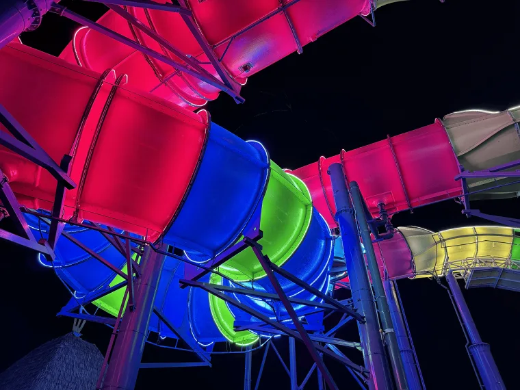 Looking up into a knot of slide tubes lit magenta, blue and green, steel supports crossing the dark sky