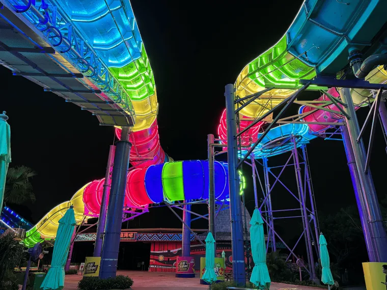 A rainbow lit tube crossing overhead above the park entrance, closed teal parasols and planting below