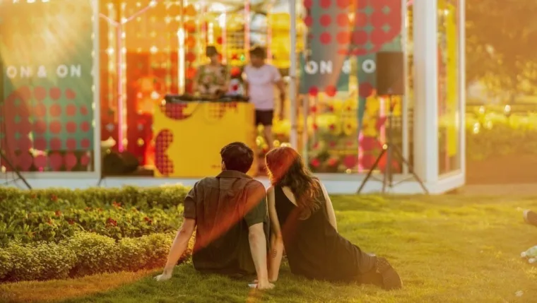 A couple sitting on the grass at sunset watching a DJ play inside the glass-walled Boom Box, patterned graphics across its windows