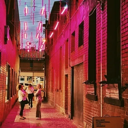Three friends walking down Hop Lane under the roof of lights, magenta wash across the brick and a Ballarat International Foto Biennale sign on the wall