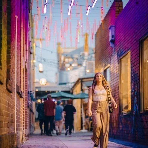 A woman in a cream outfit walking toward the camera in Hop Lane at dusk, a crowd under umbrellas behind her and coloured tubes strung overhead