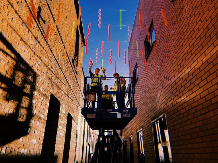 Two installers in hi-vis on a scissor lift raised between the brick walls of Hop Lane, fitting coloured perspex strips against a blue sky
