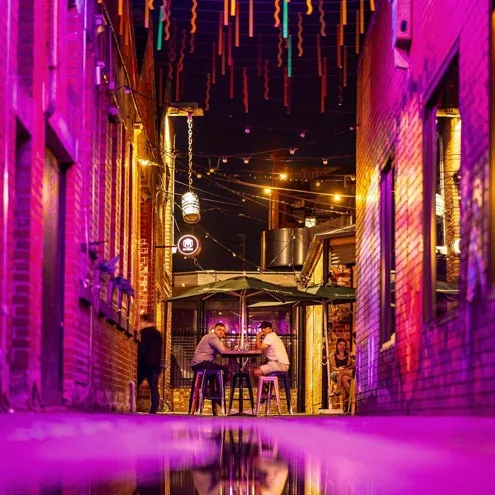 Hop Lane looking through to the lit bar frontage, drinkers seated mid-laneway under the pink roof of lights, the wet floor doubling the scene
