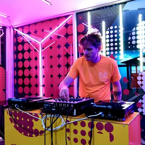 Wider view of the same DJ set, the decks on a yellow polka-dot plinth with the lit glass wall and the street visible behind