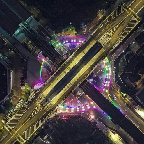 Wider overhead frame of the same junction, rooftops and side streets visible around the glowing walkway curves