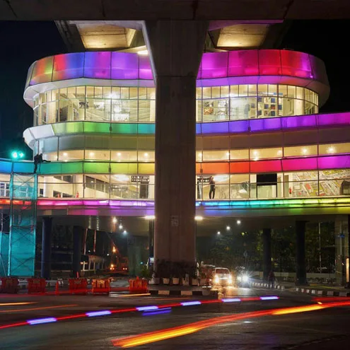 Ground level view of the round glazed concourse lit in bands of rainbow colour, car light trails across the road in front