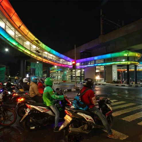 Motorcyclists waiting at a crossing under the curved walkway, its underside washed in orange, green and blue