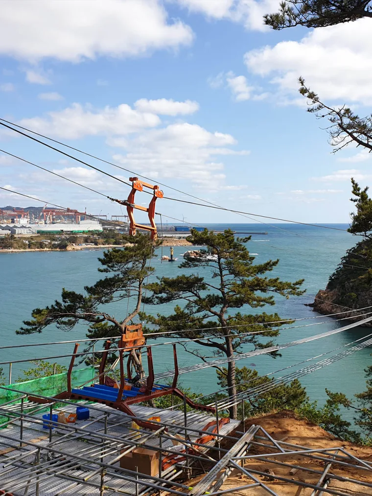 Construction shot from the deck, a worker high on the steel pylon among the stay cables above a wooded coastal inlet