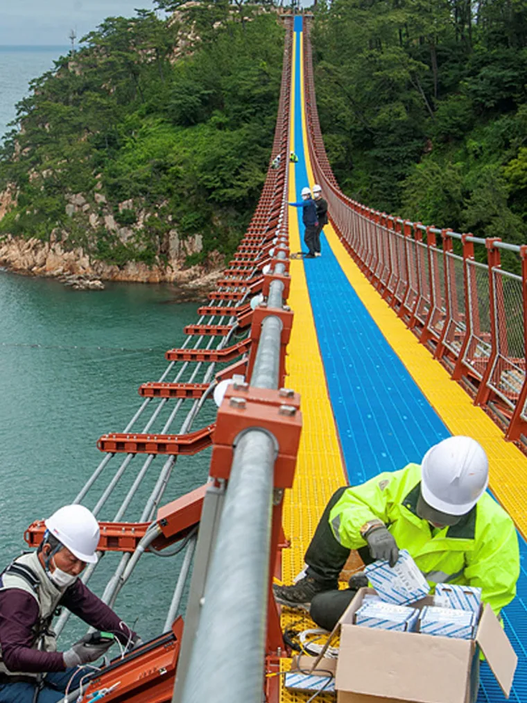 Two workers in white hard hats and hi vis kneeling on the deck fitting fixtures from a carton, green headland behind