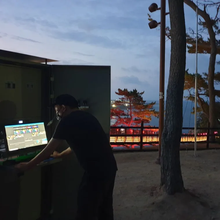 A technician at the control cabinet with a monitor at dusk, the bridge glowing red and amber through the pines behind him