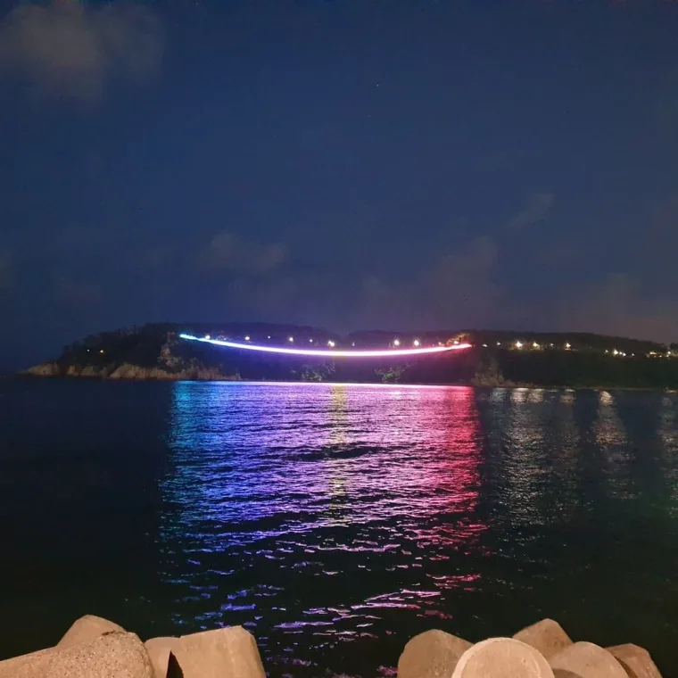 The span at night from across the water, a bright arc over the headland with magenta and blue reflections and tetrapods in the foreground