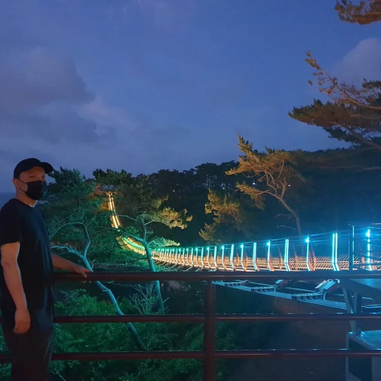 A man in a cap and face mask leaning on a viewing railing at dusk, watching the span lit in blue and orange