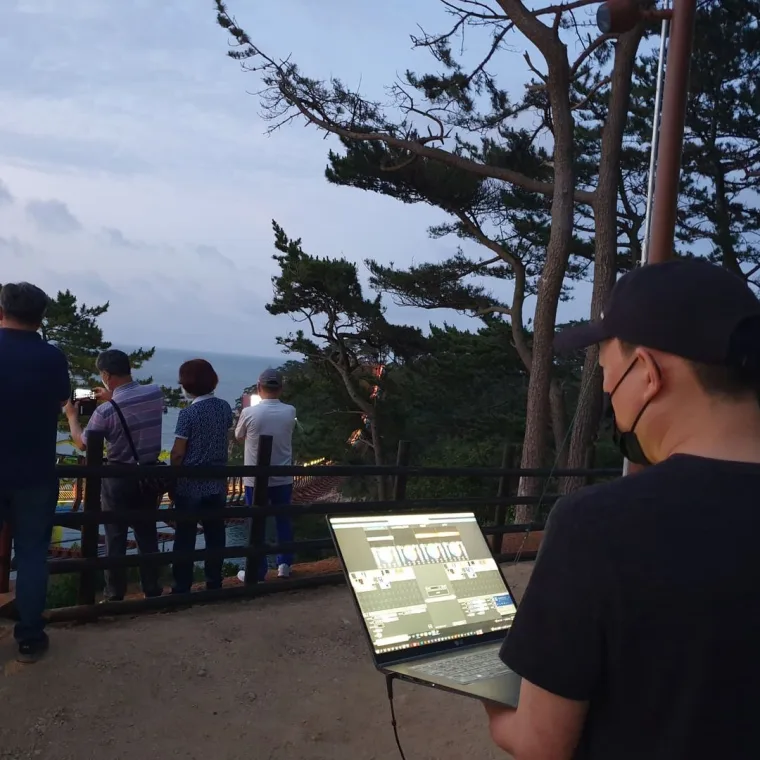 Over the shoulder of a technician with a laptop of lighting software open, onlookers gathered at the viewing rail at dusk