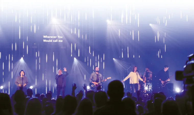 The band lit cool blue and white, vertical pixel strips and beam fans behind, song lyrics projected across the wall