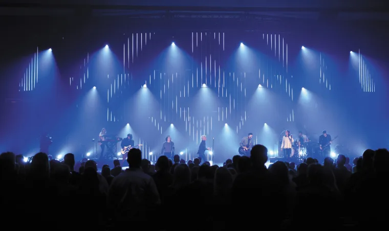 Wide blue stage with a fan of beams from above, the band in silhouette and the crowd's heads filling the foreground