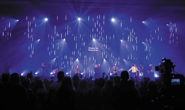 Blue and purple stage from further back, a camera operator at the right edge and lyrics floating above the players