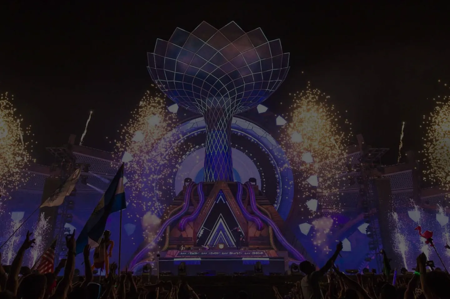 The Tree of Imagination sculpture at EDC Las Vegas lit blue and violet above the stage, fireworks arcing behind the crowd