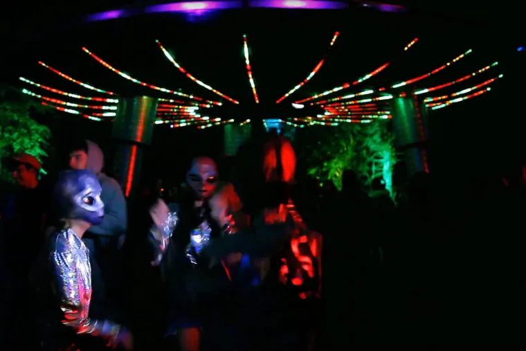 Dancers under the saucer at night, red and green pixel lines radiating overhead from its centre