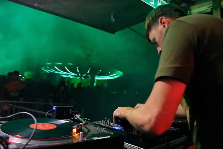 A DJ leaning over the decks in green haze, a record spinning in the foreground and the lit UFO behind the crowd