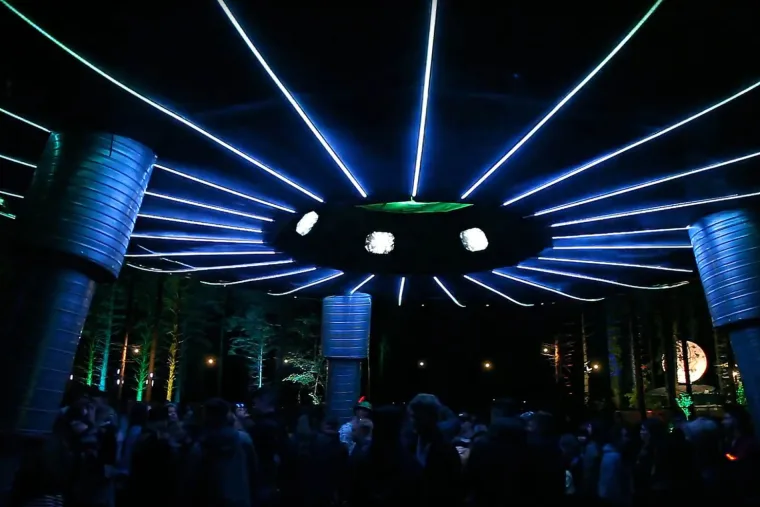 Looking up into the underside of the ship, blue spokes running out from the middle above speaker stacks and a packed dance floor