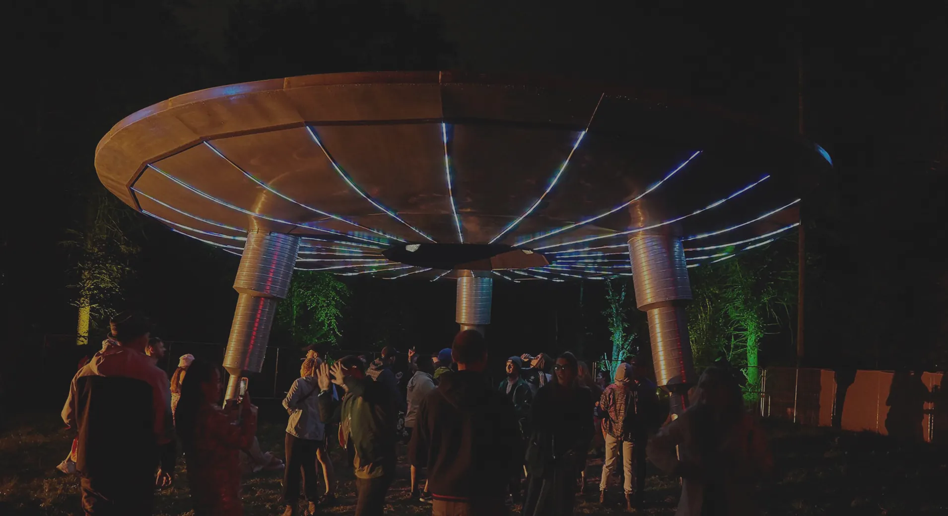 The Mothership UFO after dark with a crowd gathered beneath it, white spokes fanning across its amber underside and the three chrome legs washed in pink and blue