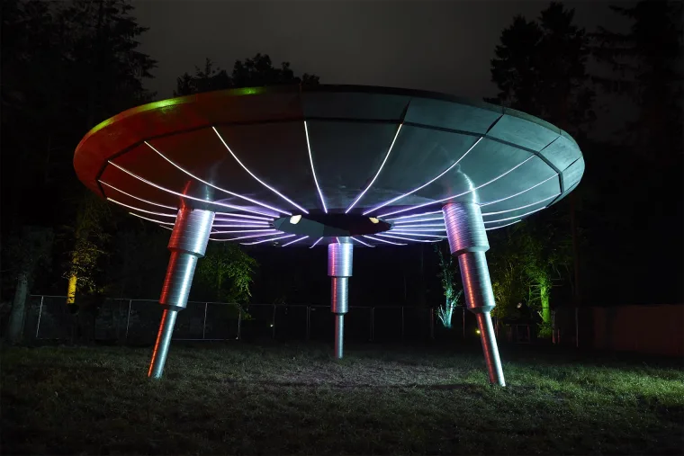 The sculpture from the front at night, the saucer rim washing from green through red with pixel lines sweeping in under the hull