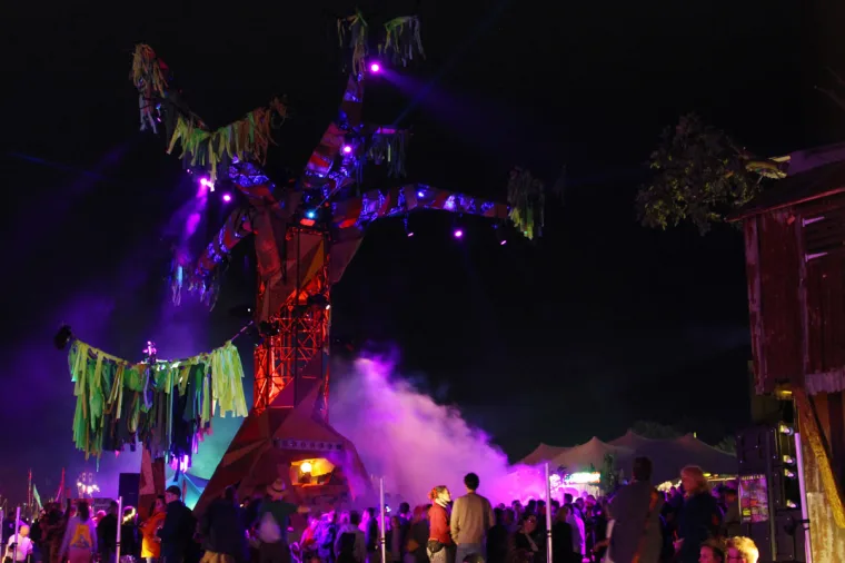Wider view of the sculpture, smoke drifting through the crowd toward the lit marquees and a timber shed at the edge of the field