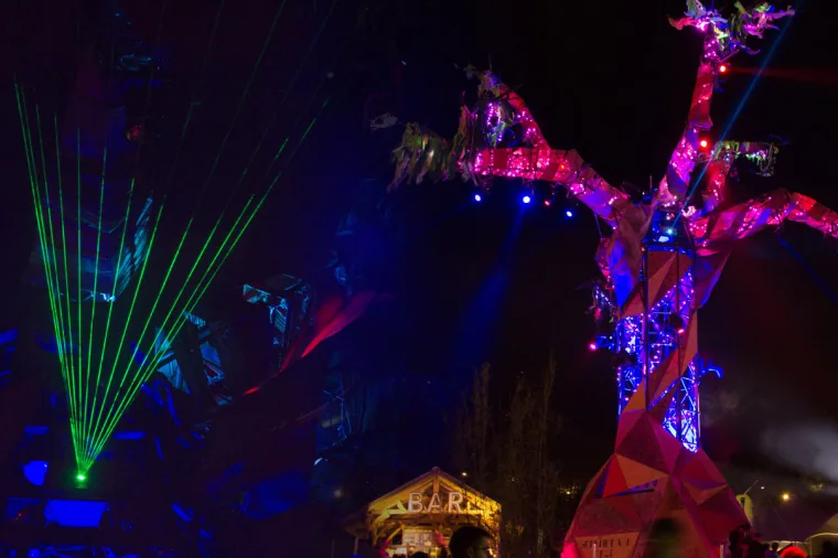 Green lasers fanning out beside the sculpture, a bar sign glowing beneath the branches
